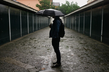 Man in casual wear and backpack standing in garage alley with umbrella in rainy autumn weather....