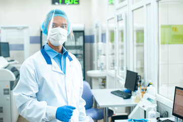 A medical worker, a laboratory assistant in a protective mask, visor, disposable cap and rubber gloves stands in a laboratory, hospital.