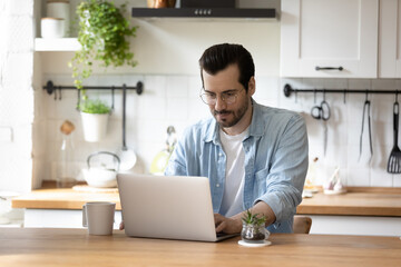 Fototapeta premium Young Caucasian male in glasses sit in kitchen work freelance on modern laptop at home, focused millennial man look at computer screen, browse surf internet on gadget, technology concept