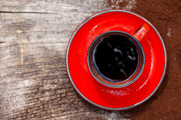 Kawa w czerwonej filiżance ze spodkiem, stojąca na drewnianej poszarzałej desce z rozsypaną, zmieloną kawą. Ujęcie z góry. - obrazy, fototapety, plakaty