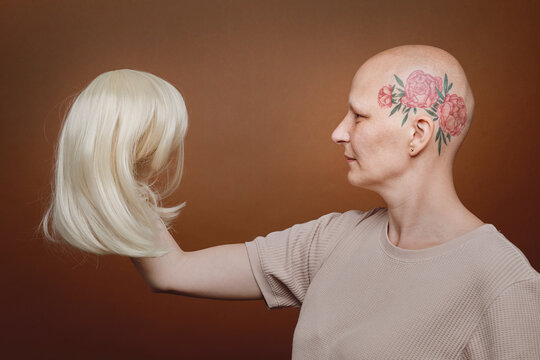 Side view portrait of confident bald woman holding wig of blonde hair against brown background in studio, alopecia and cancer awareness, copy space