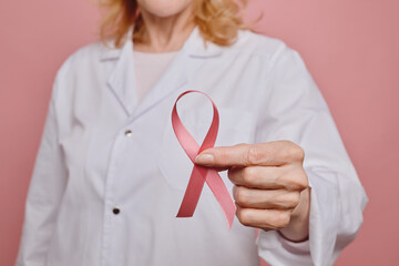 Close up of female doctor holding pink ribbon to camera while while posing against pink background in studio , breast cancer awareness concept, copy space