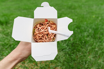Takeaway paper box with tasty wok noodles meal against green grass background. Street food and lunch concept
