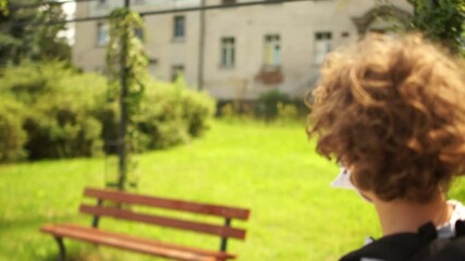Masked boy leaves school and sits on a bench in the schoolyard. Removes the mask and dishes freely. Quarantine during COVID-19 coronavirus pandemic, back to school after quarantine, social distance - Powered by Adobe