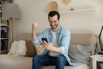 Excited millennial Caucasian male sit on couch at home triumph get pleasant good message on...