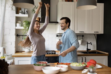 Overjoyed young Caucasian couple dancing singing preparing healthy food together in kitchen, happy millennial husband and wife have fun preparing food enjoy lazy leisure family weekend at home