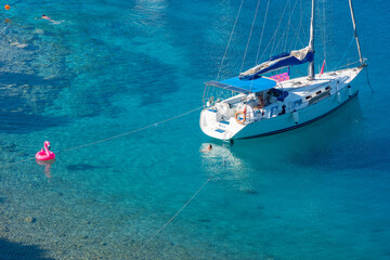 Sailing boat anchored on a beautiful blue water. People and their pink flamingo are swimming.