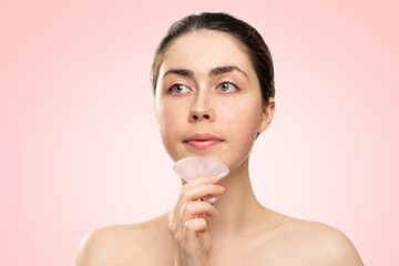 Gua Sha Massage. A woman in a white bath towel massages her chin with a quartz guasha scraper. Pink background. Copy space. Concept of alternative medicine and cosmetology