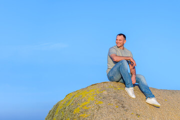 Naklejka na ściany i meble young athletic attractive man sitting on a rock against a blue sky background and smiling looking to the side with copy space background for your content