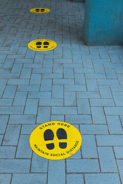 Social distancing sign stickers plastered on a brick floor near the entrance of a mall or shopping plaza. Stickers are spaced apart at recommended distance.