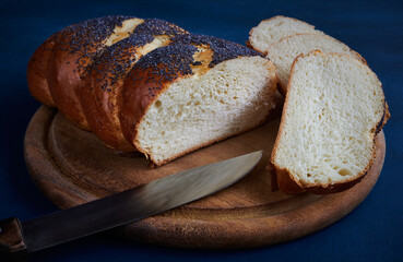 Fresh white bread loaf sprinkled with poppy on a chopping board with sliced slices and a knife, blue background