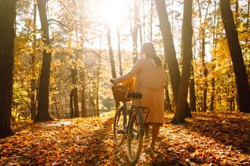 Young smiling girl with a bicycle walks in the autumn forest at sunset.
