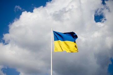 Blue and yellow Ukrainian national flag on a flagpole against a blue sky and white clouds, flag waving in the wind