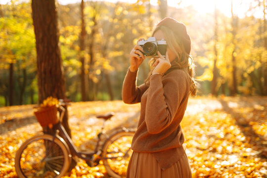 Beautiful young woman takes photos with a retro camera in autumn forest. Smiling girl enjoying autumn weather. Rest, relaxation, lifestyle concept.