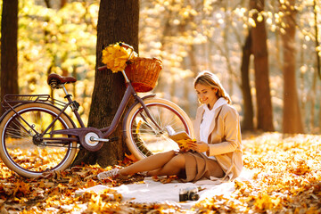 Beautiful young woman sitting on a fallen autumn leaves in a park, reading a book. Smiling girl with a bicycle enjoying rest and freedom. Relaxation and lifestyle concept.
