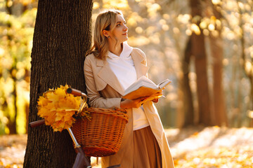Beautiful young woman sitting on a fallen autumn leaves in a park, reading a book. Smiling girl...