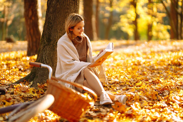 Beautiful young woman sitting on a fallen autumn leaves in a park, reading a book. Smiling girl...