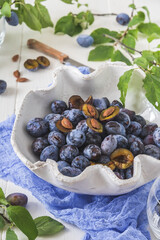 White curved bowl filled with ripe plums, some of them sliced, on a white wooden table with green...