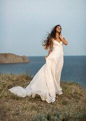 Fototapeta na wymiar An attractive and slender bride in fashion wedding dress gently touches her face and dreams with her eyes closed against the background of the bay. The bride's dress and hair flow in the wind.