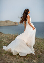 Fototapeta na wymiar A tanned bride with her wedding dress fluttering in the wind looks out at the horizon, against the background of the Bay and hills at dusk.