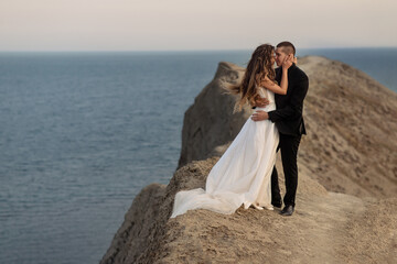 Fototapeta na wymiar The bride and groom gently embrace each other standing at the edge of the cliff. Bride's hair and her beautiful wedding dress flutter in the wind.