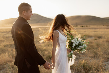 Happy bride and groom hold hands in the autumn field on the background of sunset hills and...