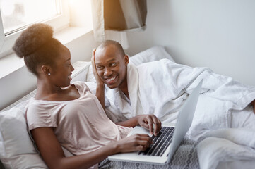 handsome african man supports his wife while she is working at home, woman sit with laptop on bed, man lie next to her and smile