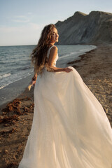 Fototapeta na wymiar Attractive and happy bride in a fluttering luxury wedding gown with a big hem runs along the beach against mountains in the sunset light.
