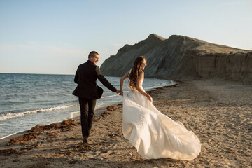 Fototapeta na wymiar Beautiful wedding couple run along the beach near the coastline holding hands against the background of rocks in the sea. Smiling bride runs along the sand with a luxurious flowing hem of her dress.