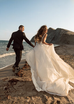 Happy wedding couple run along the beach near the coastline holding hands against the background of rocks in the sea. A beautiful and smiling bride runs along the sand with a flowing hem of her dress.