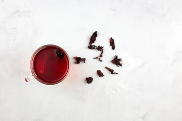 A glass cup with red  hot hibiscus tea or sudanese rose on light grey background with dry tea leaves. Top view.  Copy space.  