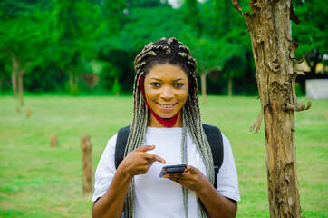 Naklejka premium young beautiful african student wearing face mask preventing herself from the outbreak in the society feeling excited about what she saw on her cellphone and points to her phone