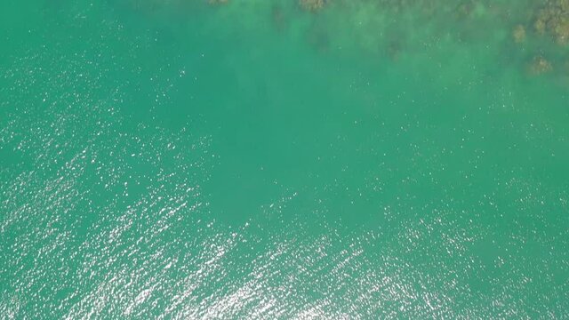 Directly above the Malaysian coast at the Blue Lagoon and Cape Rachado lighthouse. Further out of the beach, the turquoise blue sea at the stony cliff. People swim in the shallow water of the lagoon.