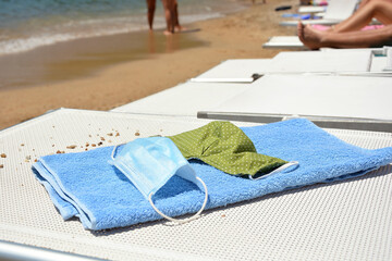 A protective mask made of fabric and a medical mask lie on a towel on a white lounger on the beach at sea.  The concept of a seaside vacation after quarantine due to the COVID 2019 Coronavirus.