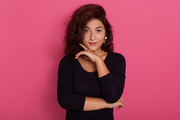 Photo of lovely female keeps hand under chin, posing with good emotions, wearing black shirt, standing isolated over pink background, stylish lady with make up.