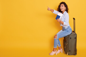 Smiling girl in stylish clothing isolated over pink background. Passenger traveling abroad, has air flight journey, holding passport and ticket while sitting on suitcase, pointing index finger aside.