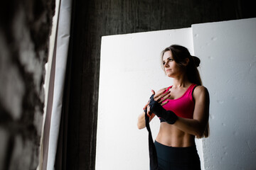 Obraz premium Portrait of a young woman kickboxer in a pink top who ties a black elastic bandage on her hand before fighting and training on a white background.