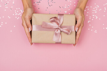 Women hands holding a gift or gift box decorated with confetti on a pink pastel table top view