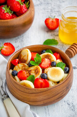 Pancake cereal, mini pancakes in a wooden bowl with fresh strawberries, blueberries and honey. Trend food. Vertical, copy space.