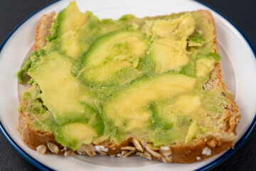 Cereal bread and ripe avocado sandwich on a white plate with a blue edge on a dark stone surface