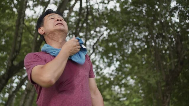 Asian middle age man wiping sweat on his face and neck during exercise at the park, standing under trees and rest after exercising, body wellness tired exhausted, medical health condition, low angle