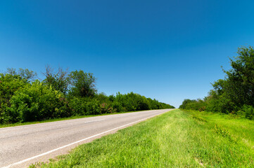 Fototapeta na wymiar Country asphalt road in summer with green grass and trees on the roadsides against the blue sky. Sunny day background for transportation and transportation logistics companies