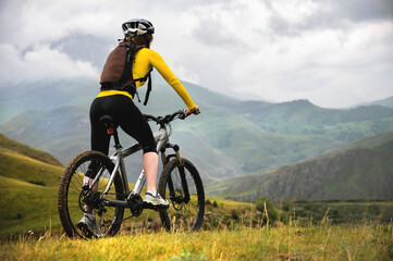 Obraz premium A young slender girl with a backpack and a bicycle helmet sits on a bicycle high in the mountains on a cloudy day and looks at the mountains and clouds.