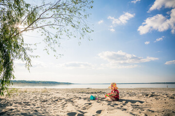A little cute girl of three years old in a red dress and a panama hat sits on the beach and plays in the sand with plastic toys: a bucket, molds and a shovel. Summertime trip. Family holiday.