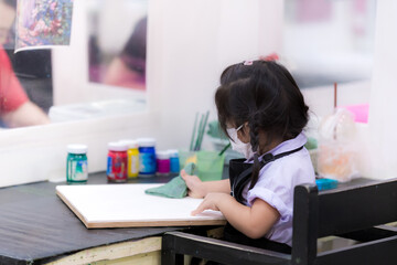 Student using a rag to wipe a canvas. Child preparing to learn water coloring by special tutor that her parents brought to study after school. Kid ware cloth face mask. Cute Asian children aged 3 year