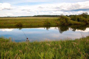 A fisherman catches fish sitting on the Bank, against the background of a rural landscape with a river on a cloudy day. The river reflects the trees and the sky with clouds.