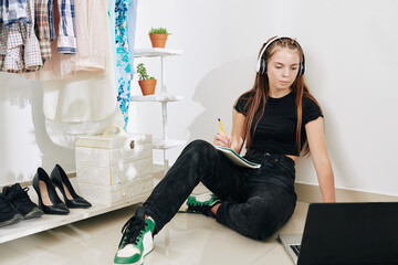 Teenage girl in headphones sitting on ther floor in her room, watching educational video on laptop and taking notes in copybook