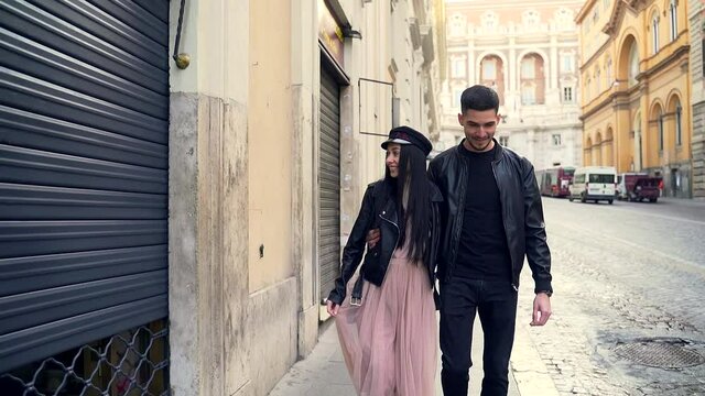 Portrait of a young couple of happy tourists walking hugging and holding hands. Stylish hipster guy and girl in black clothes, leather jacket and dress. A hat and sunglasses. go street old town