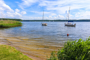 Small beach at Nidzkie lake and sailboats in distance near Ruciane Nida town, Mazury Lake District, Poland