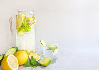 Refreshing lemonade in a tall decanter and glass on a gray background. Next to lemon, lime, cucumber and mint. Horizontal position, space for text